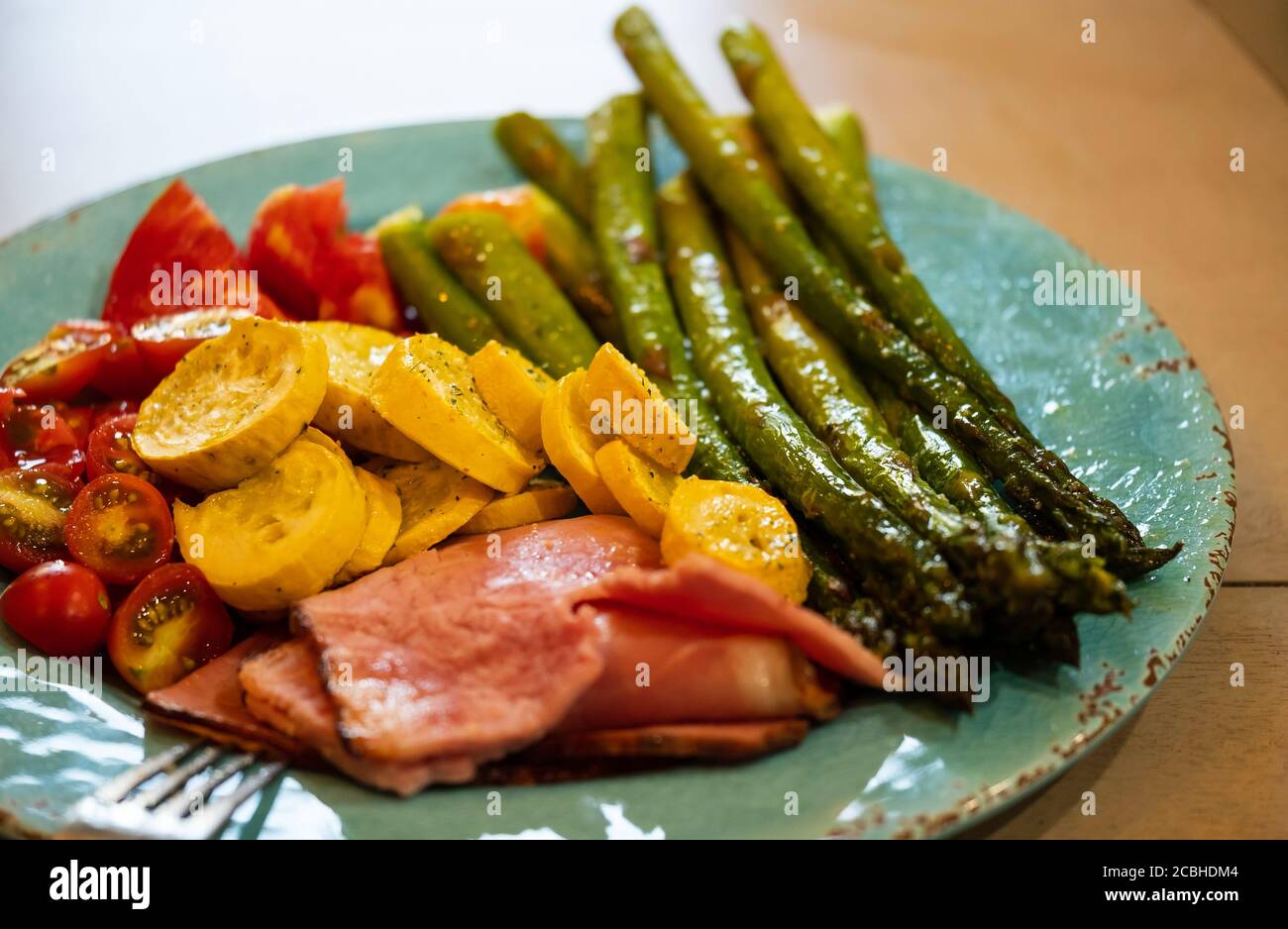 Seasoned asparagus, yellow squash, and tomatoes drizzled with olive oil and broiled. Served with ham slices on a turquoise dinner plate. Stock Photo