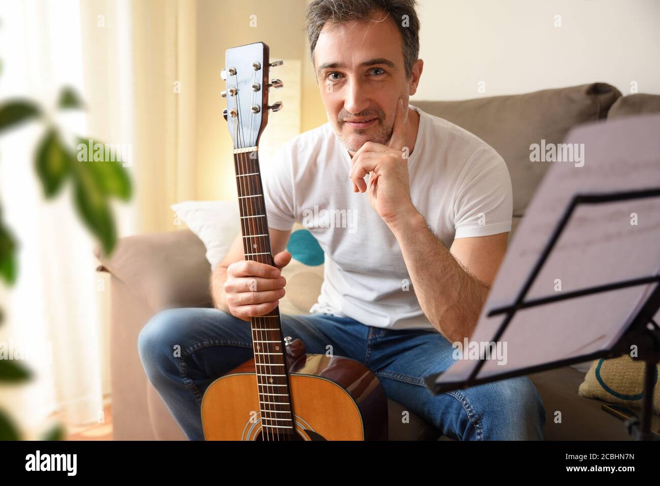 Smiling man with his acoustic guitar in hand looking straight ahead sitting on the sofa with music stand with songs at home Stock Photo