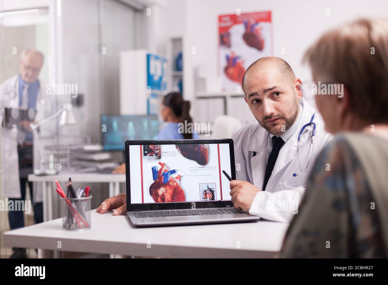 Cardiologist talking with senior patient in hospital office about her heart diagnosis and recovery treatment wearing white coat and stethoscope. Elderly doctor writing on clipboard in clinic corridor. Stock Photo