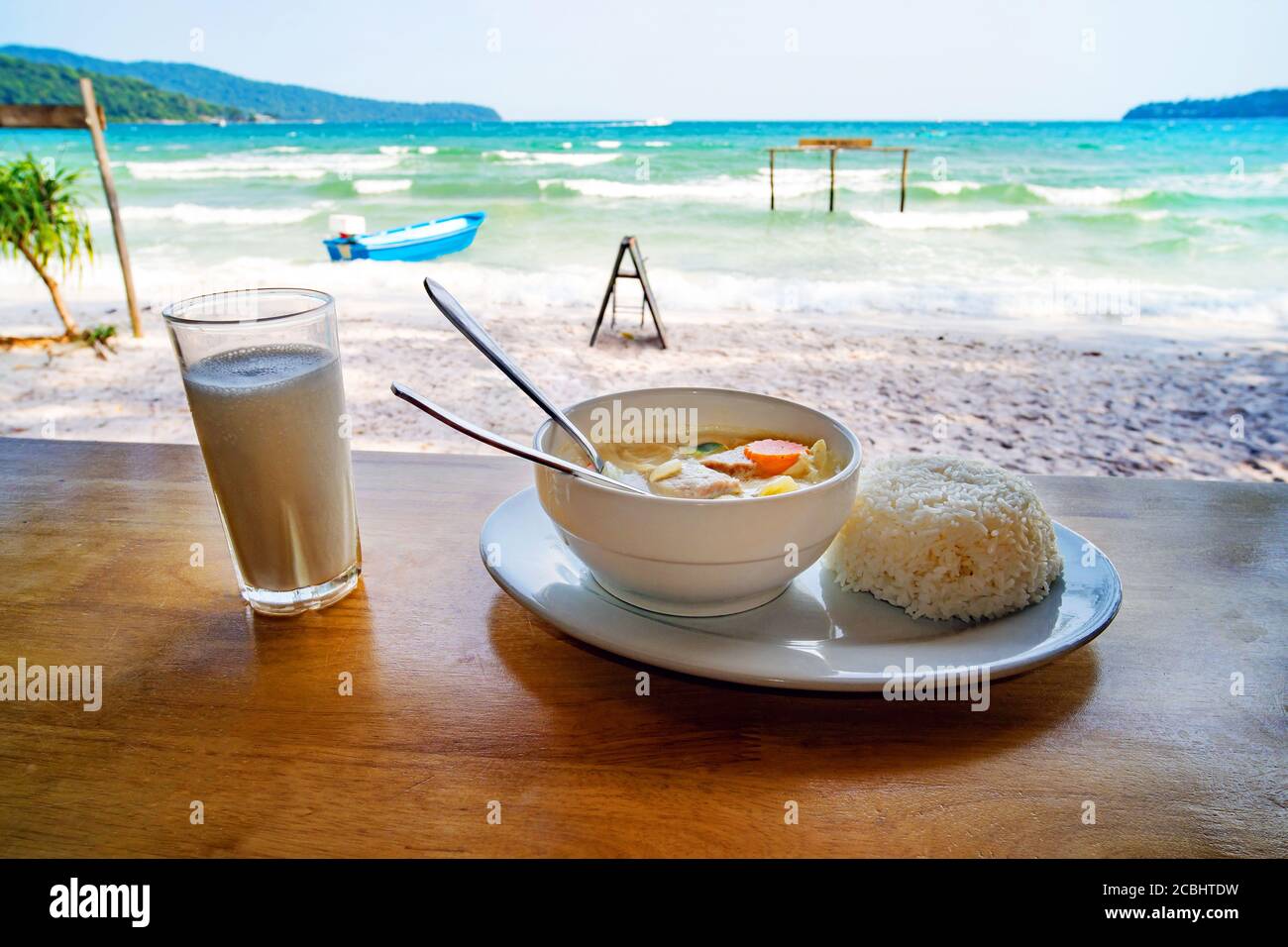 Thai cuisine. A plate of soup with coconut milk and boiled rice on the table against the beautiful sea landscape. Breakfast at the beach restaurant Stock Photo