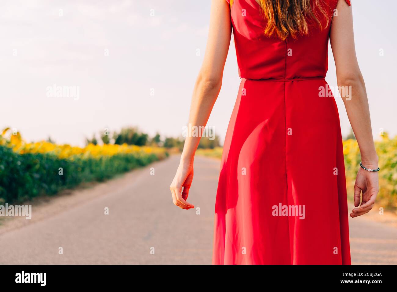 woman with red dress backwards in road with sunflowers Stock Photo