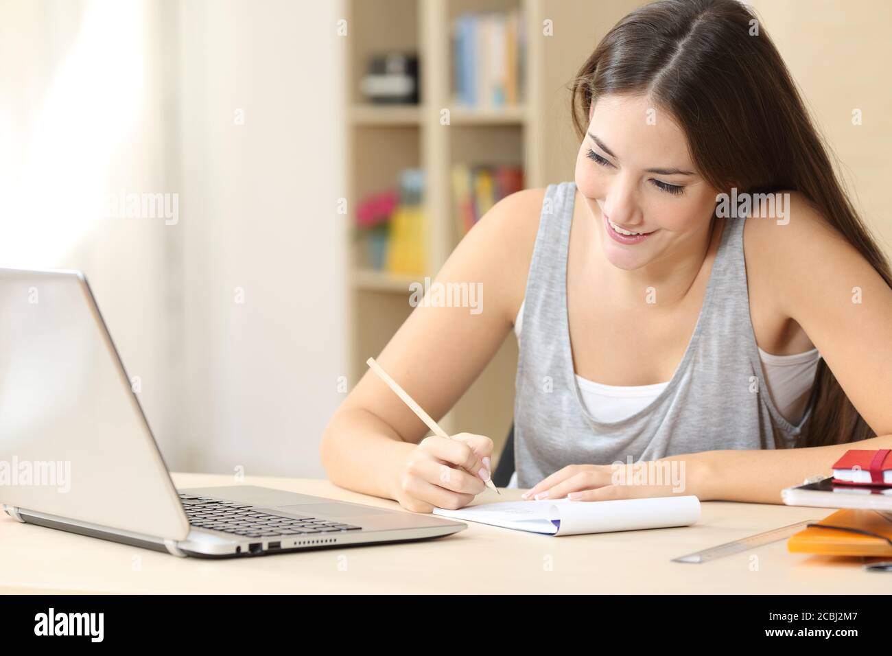 Happy student woman reading on laptop taking notes on notebook sitting on a desk at home Stock Photo