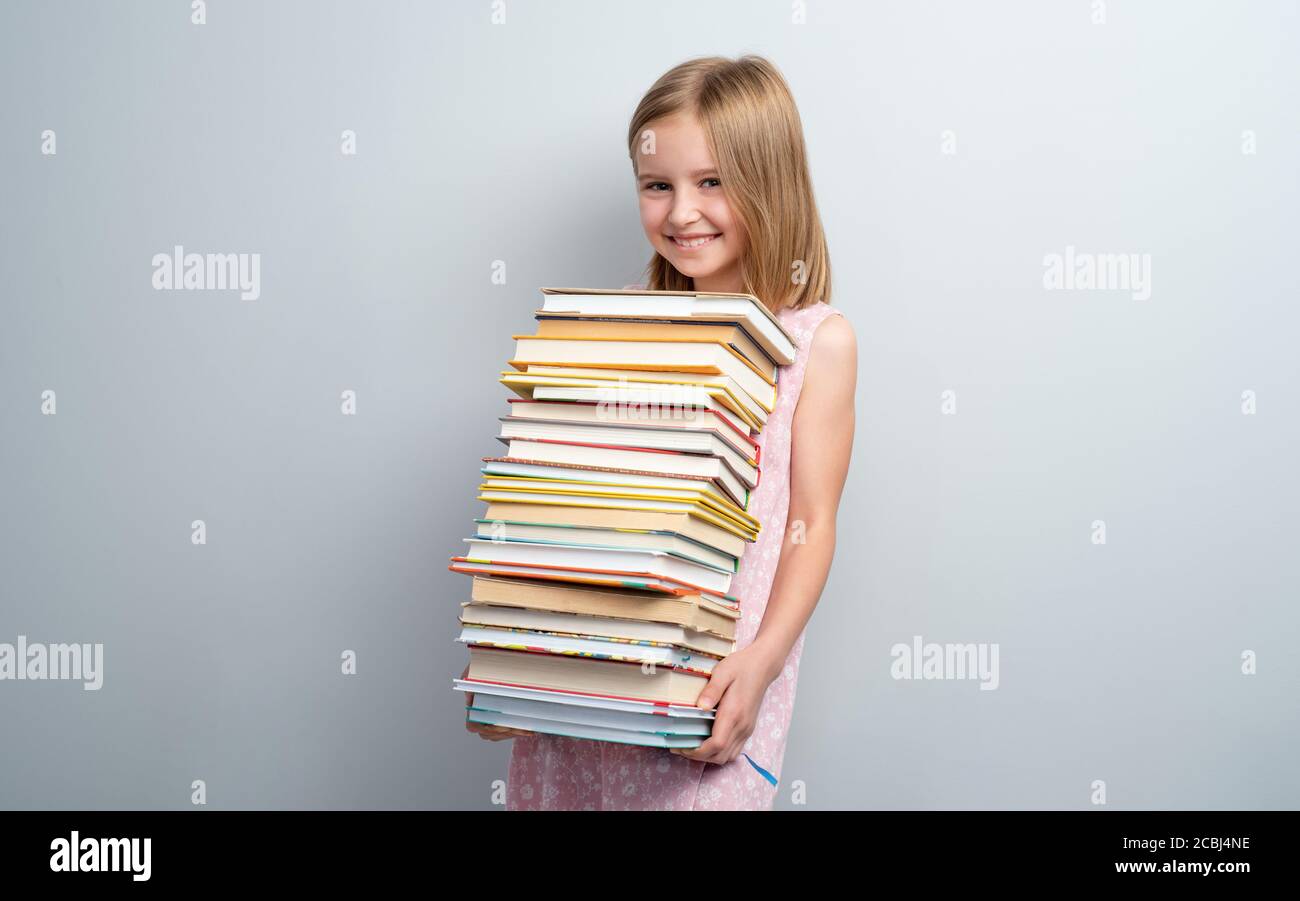Smiling school girl holding stack of books on a grey wall background ...