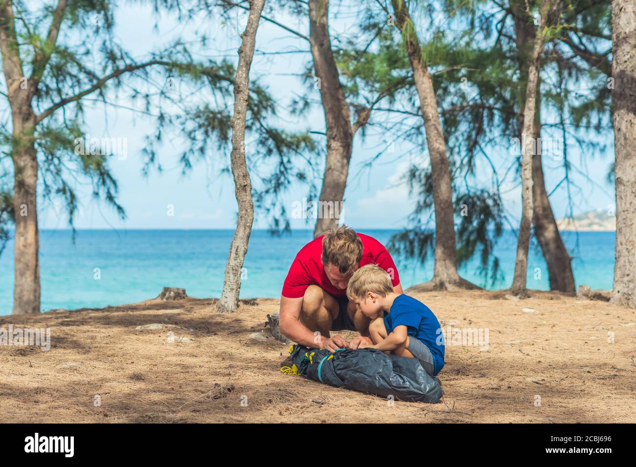 Camping people outdoor lifestyle tourists putting up setting up green grey campsite summer forest near lazur sea. Boy son helps father assembling tent Stock Photo