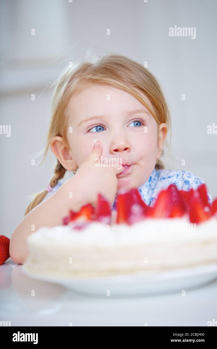 Girl secretly tastes cream from a strawberry cake in the kitchen Stock