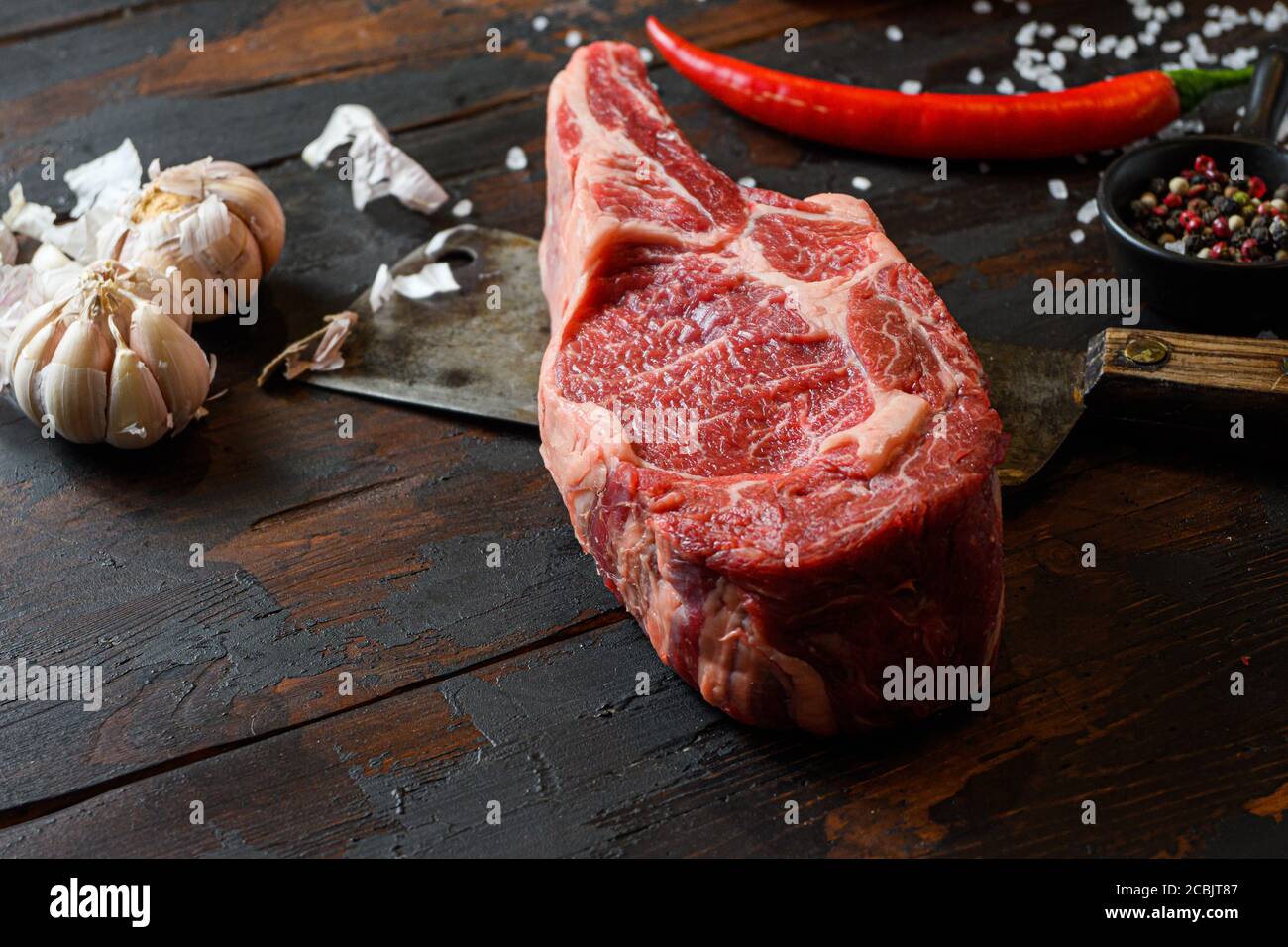 Raw dry Cowboy steak on dark wooden vintage table background. Spencer steak or beauty steak BBQ with seasonings, rosemary, tomato, chikie, garlic Stock Photo