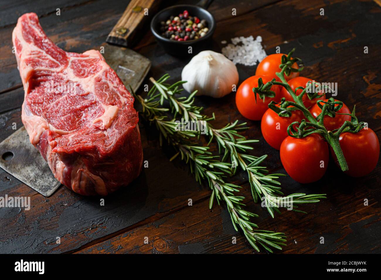 Raw Cowboy steak on meat cleaver. Organic farm marbled prime black angus beef. Dark wooden background. Side view. With seasonings, peppercorns, chilie Stock Photo