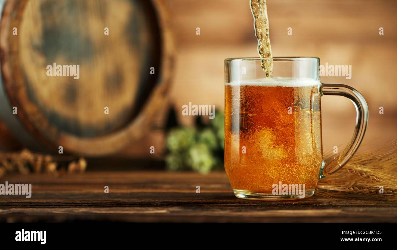 Pouring beer into glass pint, placed on wooden table with keg. Stock Photo