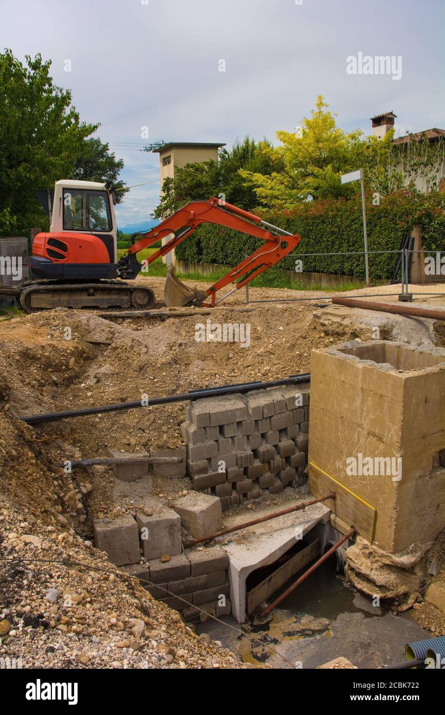 A sewer well trench & a compact crawler excavator with rotating house platform & continuous caterpillar track on a sewer replacement site in NE Italy Stock Photo