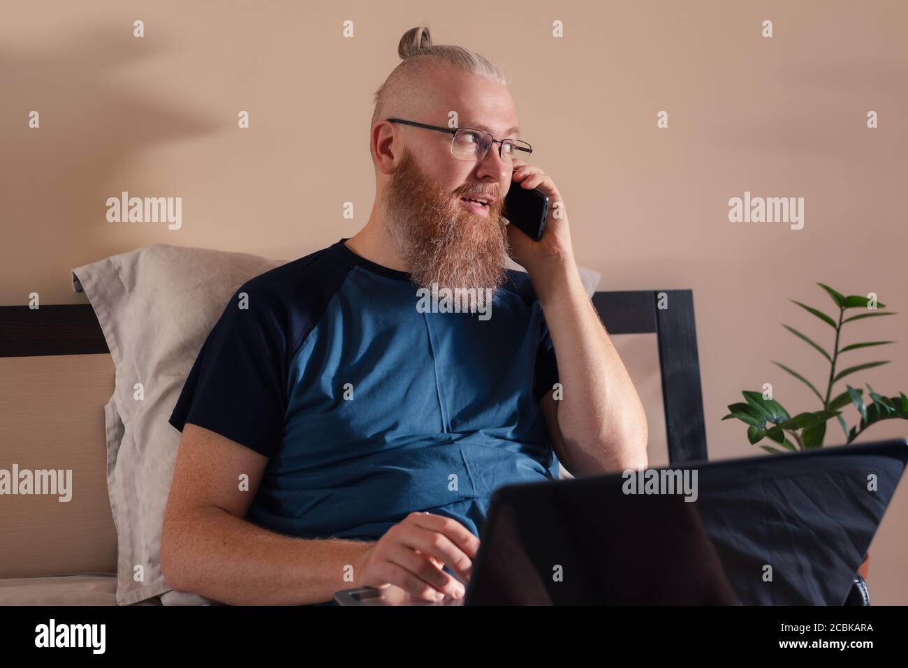 Middle aged businessman working from home talking on mobile phone while working on laptop. Smiling red bearded man solves work issues remotely Stock Photo