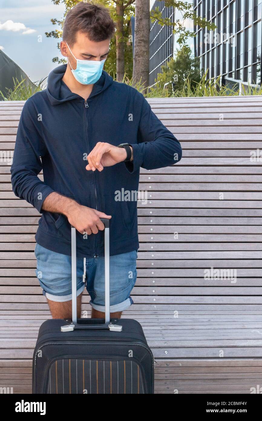 Man in protective face mask holding luggage and looking at watch on hand. Tourist wearing medical with suitcase glancing at watch adn waiting for trip Stock Photo