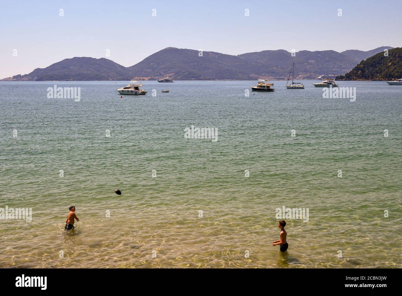Two kids playing by throwing a rugby ball on the sea shore of the Gulf of Poets with the promontory of Porto Venere, Lerici, La Spezia, Liguria, Italy Stock Photo