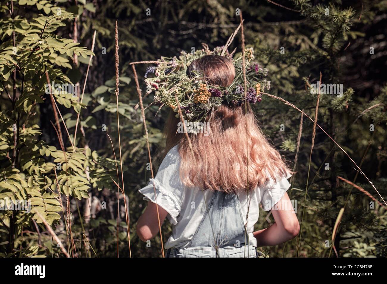 Cute girl with a wreath of forest flowers on her head in the sunlight. Back view Stock Photo