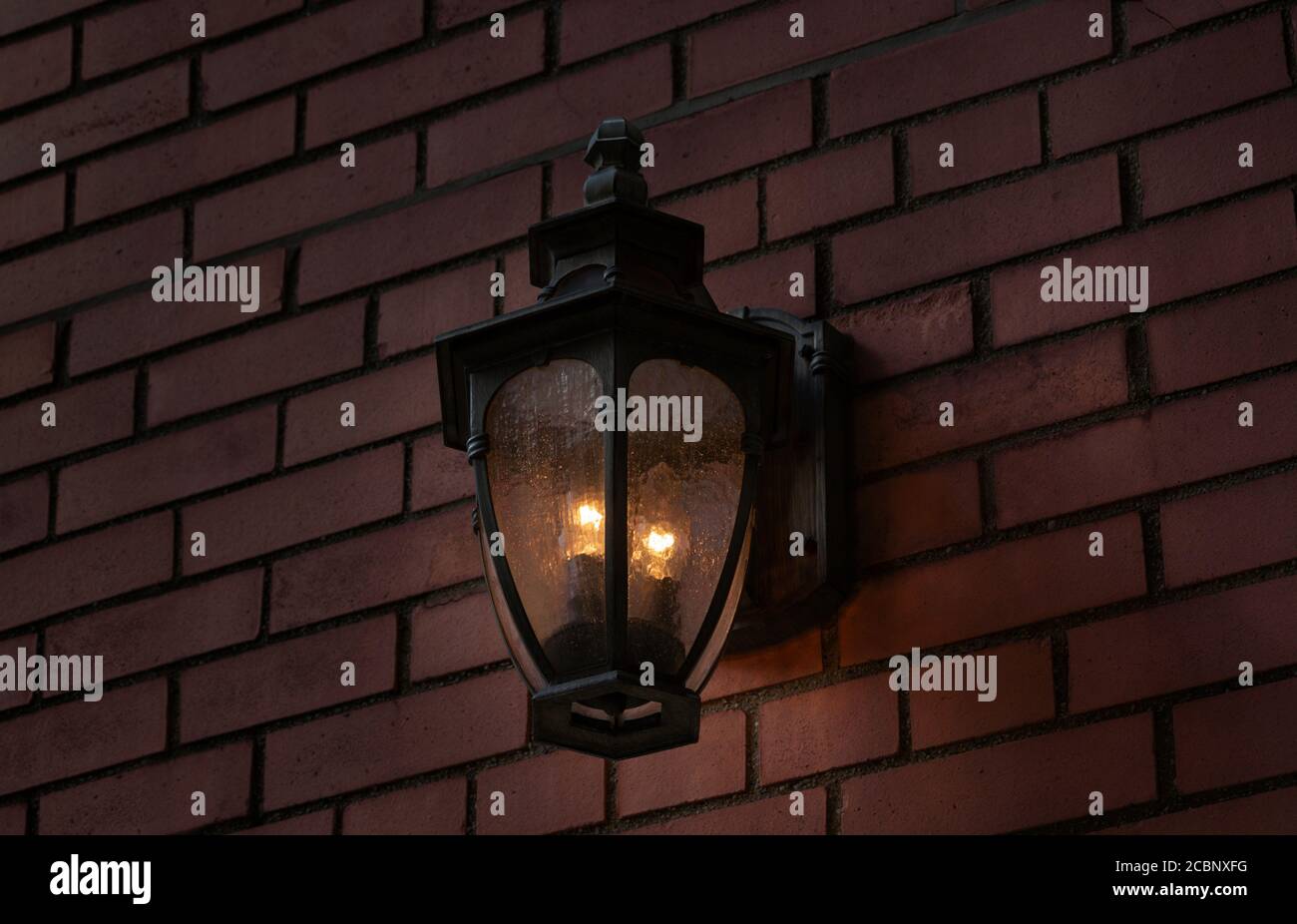 a black iron metal antique illuminated wall sconce on an exterior brick wall at night giving off a warm glowing light Stock Photo