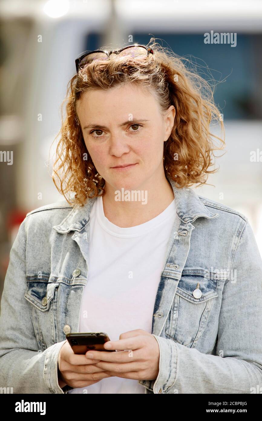 Young woman with denim jacket and sunglasses in curly hair holds smartphone in her hand, Cologne, North Rhine-Westphalia, Germany Stock Photo