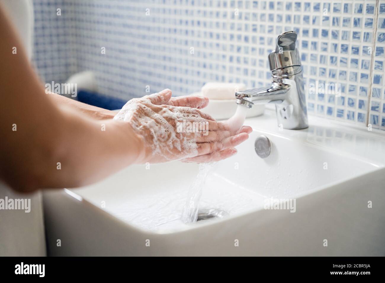 Young woman washes hands in bathroom washbasin under running tap water. Rubs hands together with soap Stock Photo