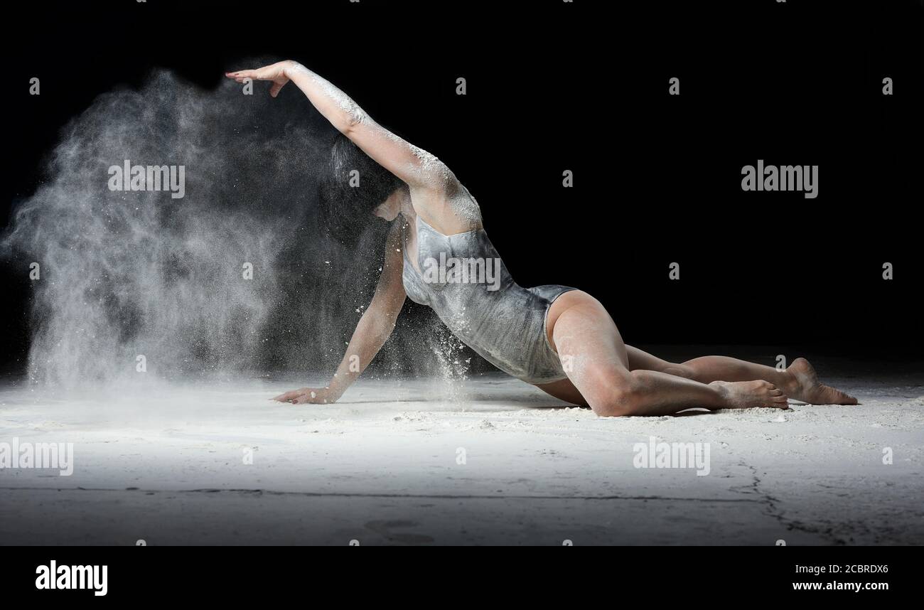 young beautiful woman with black hair is dressed in a sports black bodysuit and lies on the floor and throws white flour over her, black background Stock Photo
