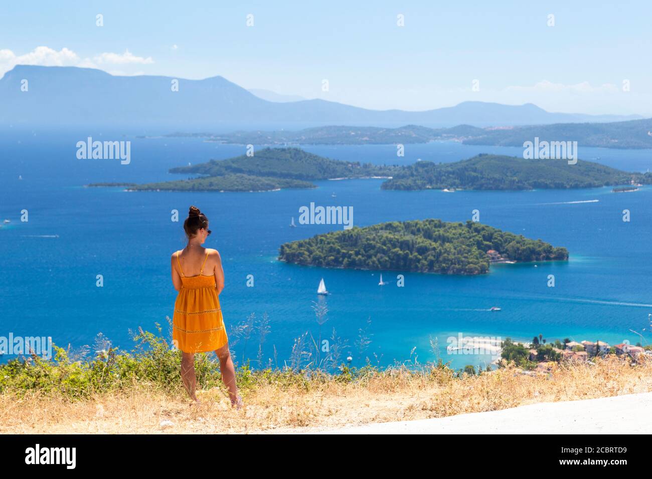 A girl in a yellow dress enjoys a coastal view from a road above Nidri