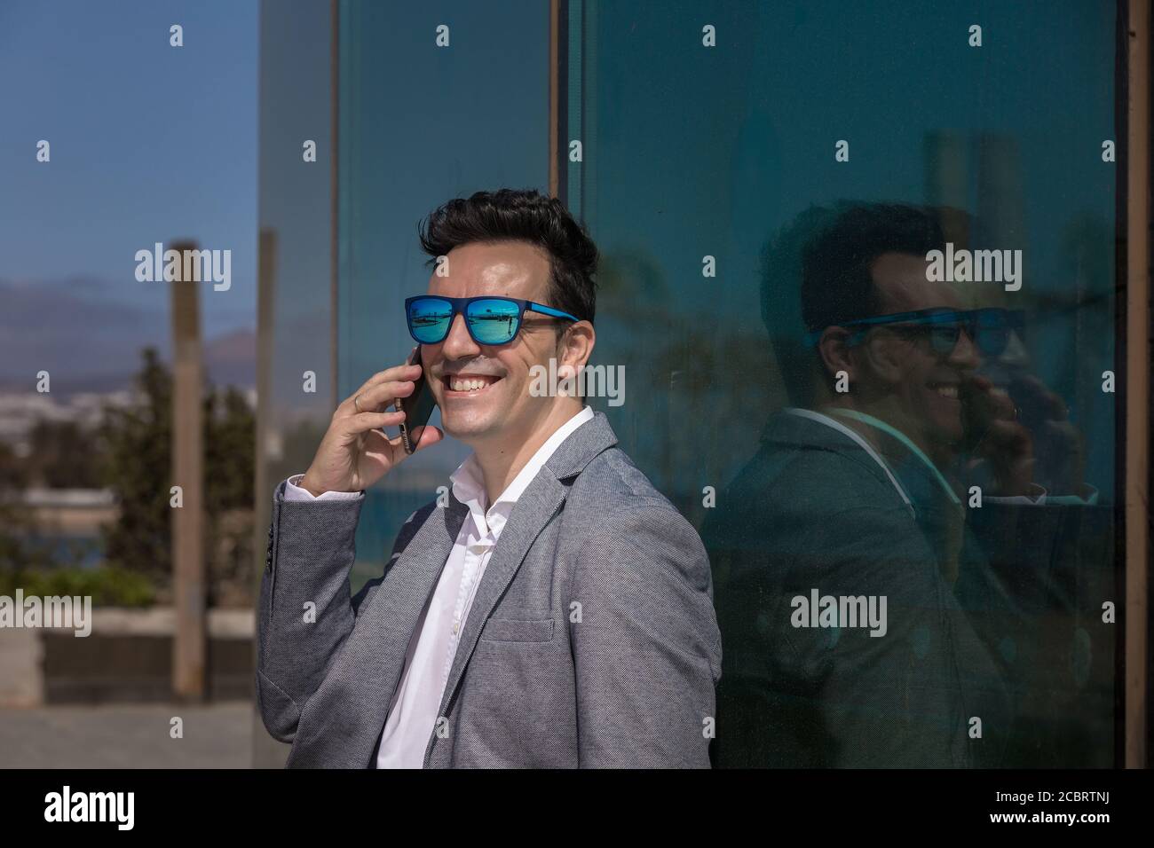 Optimistic adult businessman in sunglasses smiling and talking on smartphone while leaning on glass wall in city Stock Photo