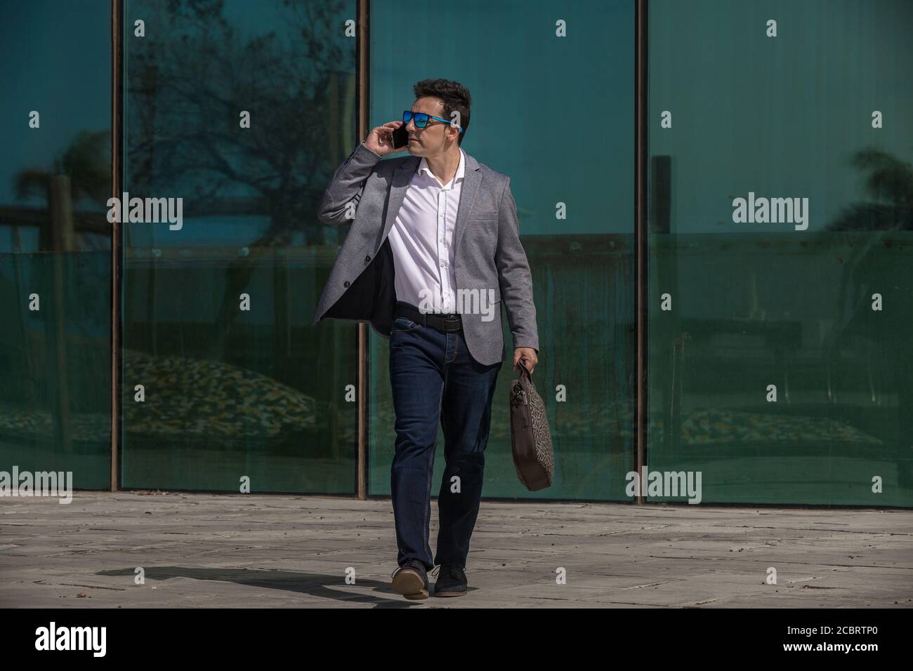 Full body adult man in smart casual clothes talking on smartphone and looking away while walking near glass building in downtown Stock Photo