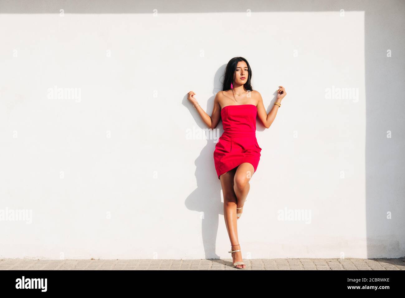 portrait of elegant latin young girl with long brunette hair and red dress on white wall with strong shadow, looking at camera. copy space Stock Photo