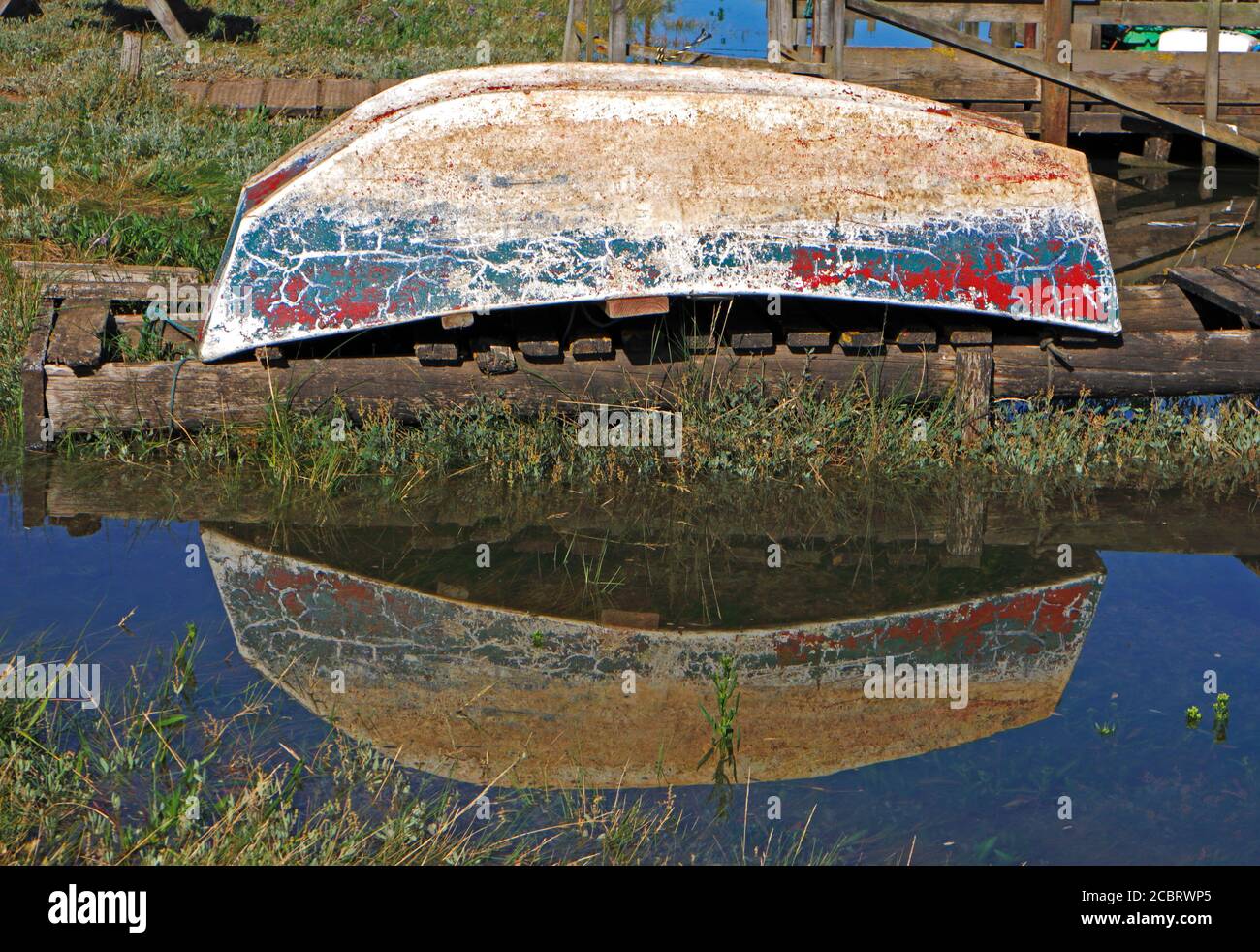 A small upturned dinghy with distressed paintwork and reflection on the quayside at the North Norfolk harbour of Thornham, Norfolk, England, UK. Stock Photo