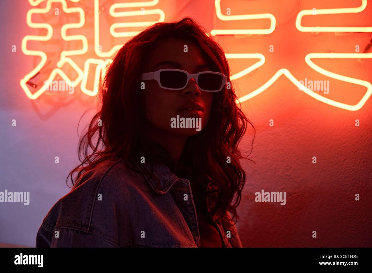 African woman wears trendy glasses stands in red neon backlight in night club. Stock Photo