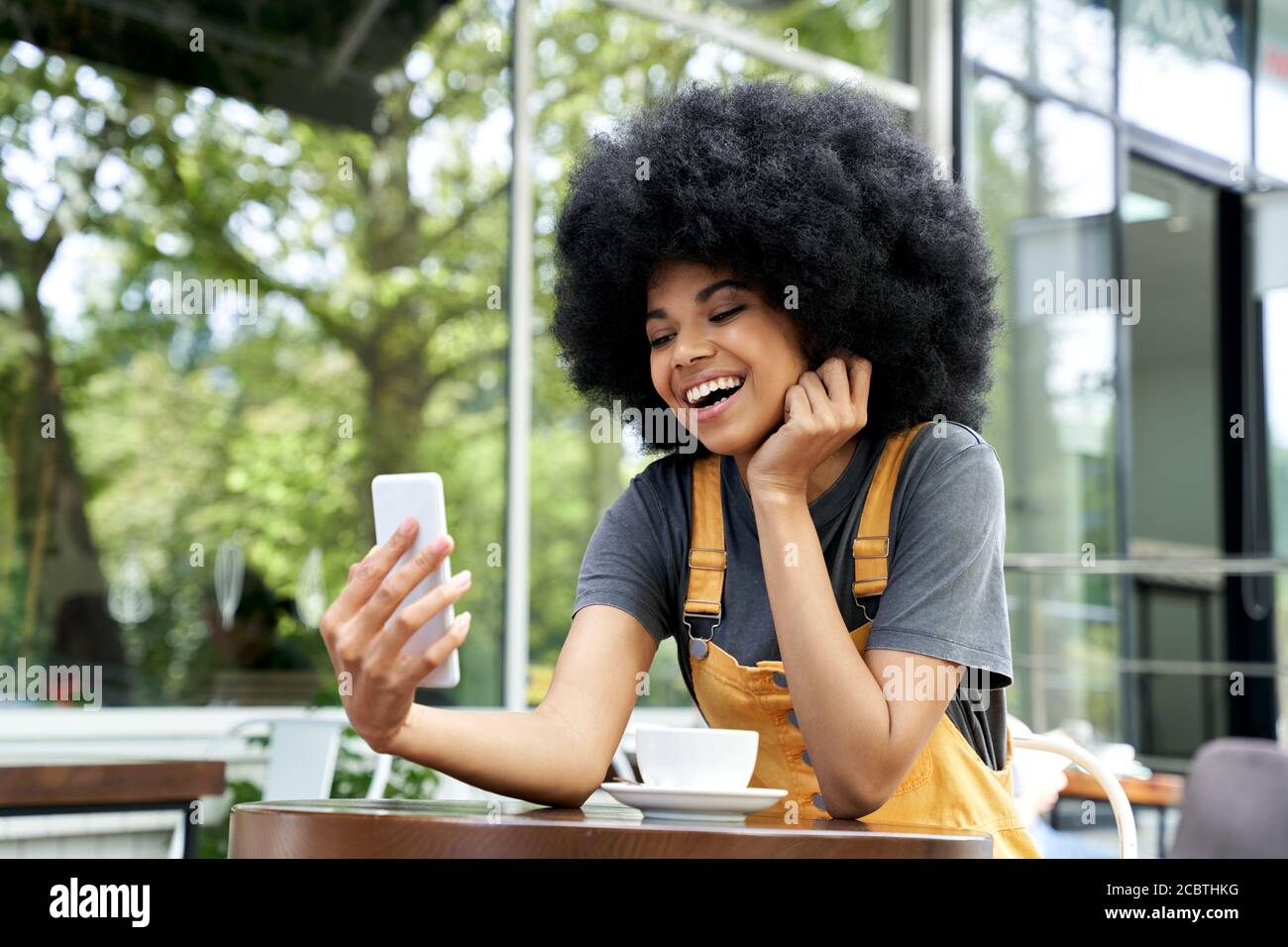 Happy African teen girl making video call on phone sits at outdoor cafe table. Stock Photo