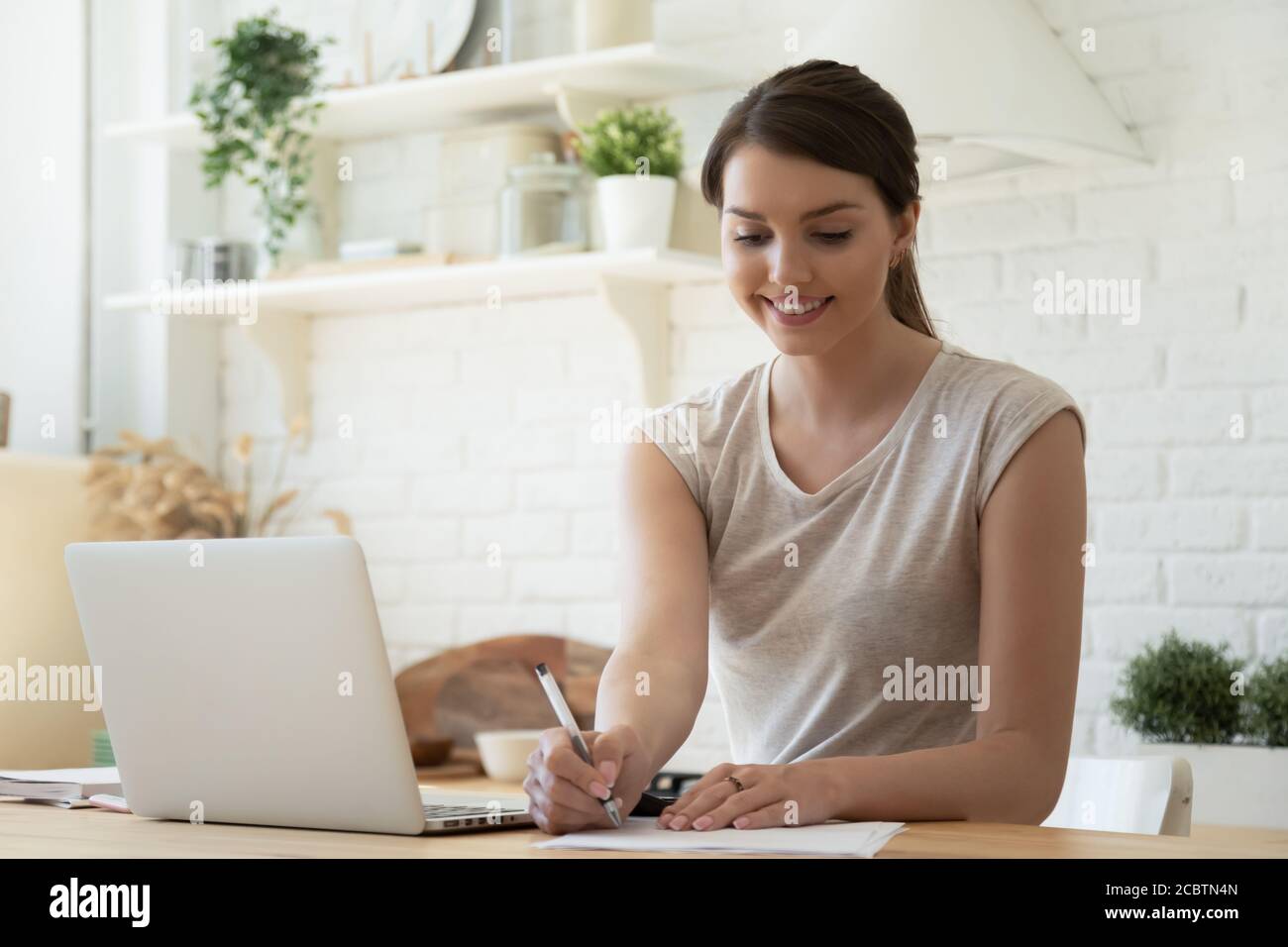 Smiling female working with laptop and making notes at kitchen Stock Photo