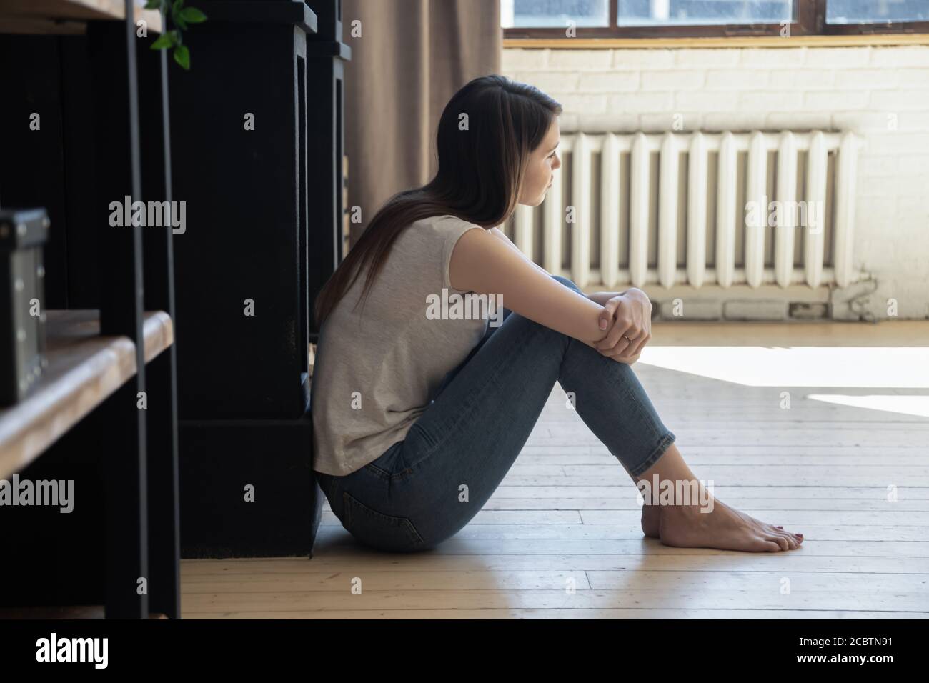 Thoughtful depressed young female sitting on floor hugging knees Stock Photo
