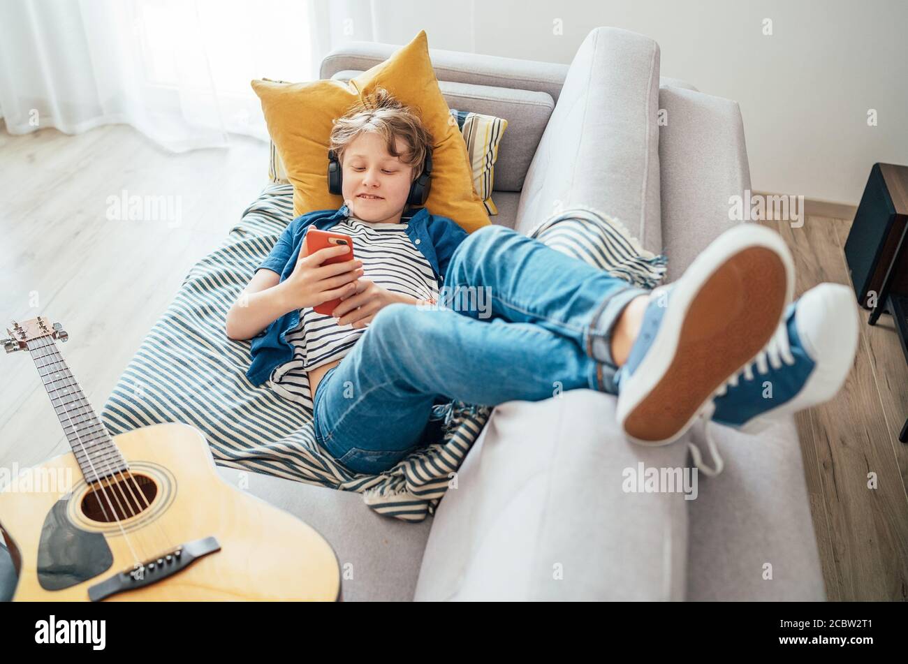 Preteen boy lying with guitar on cozy sofa dressed casual jeans and new sneakers listening to music and chatting using wireless headphones connected w Stock Photo