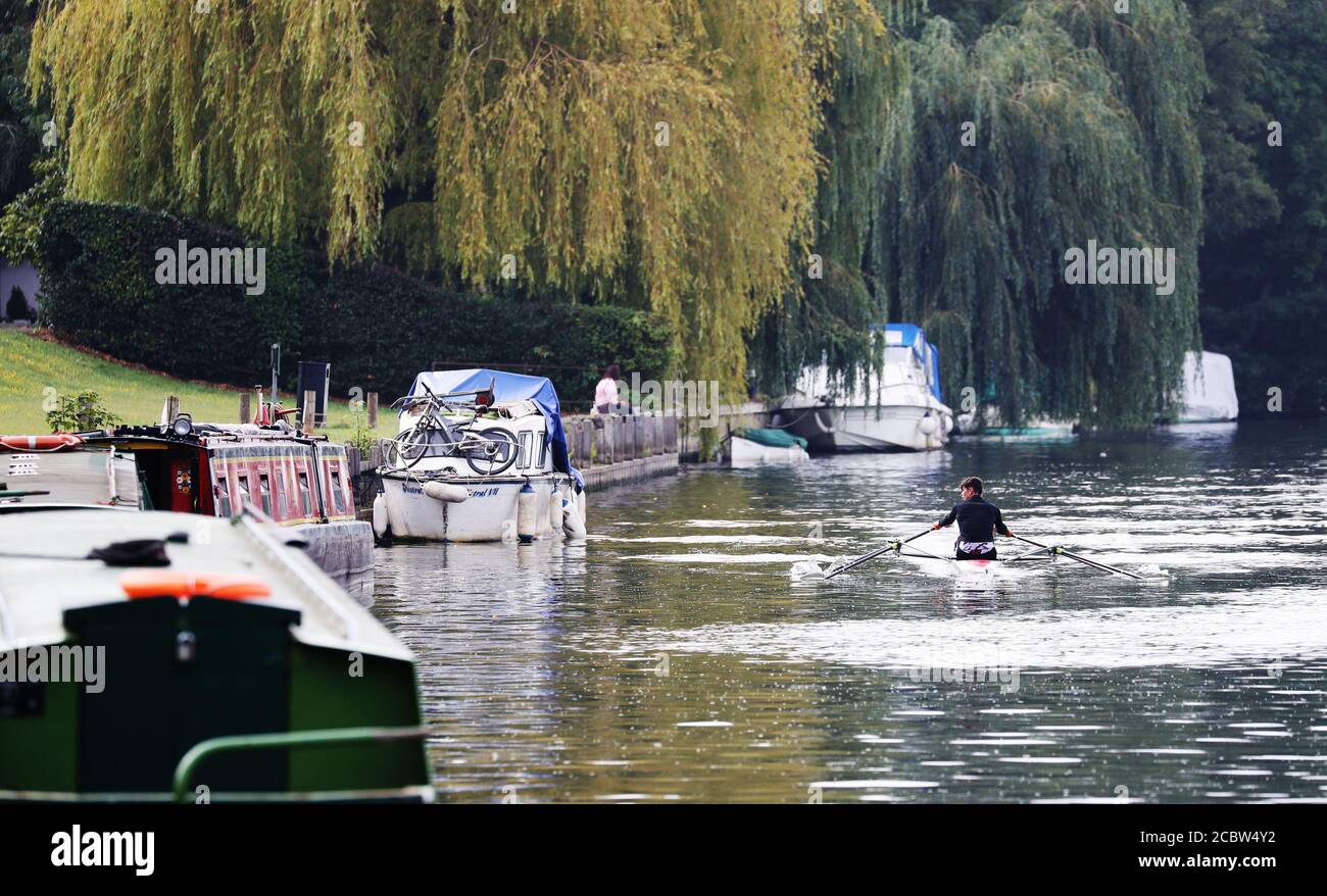 Rowers pass pleasure craft and long boats moored along the banks of the river Thames near Bray, Berkshire. Stock Photo