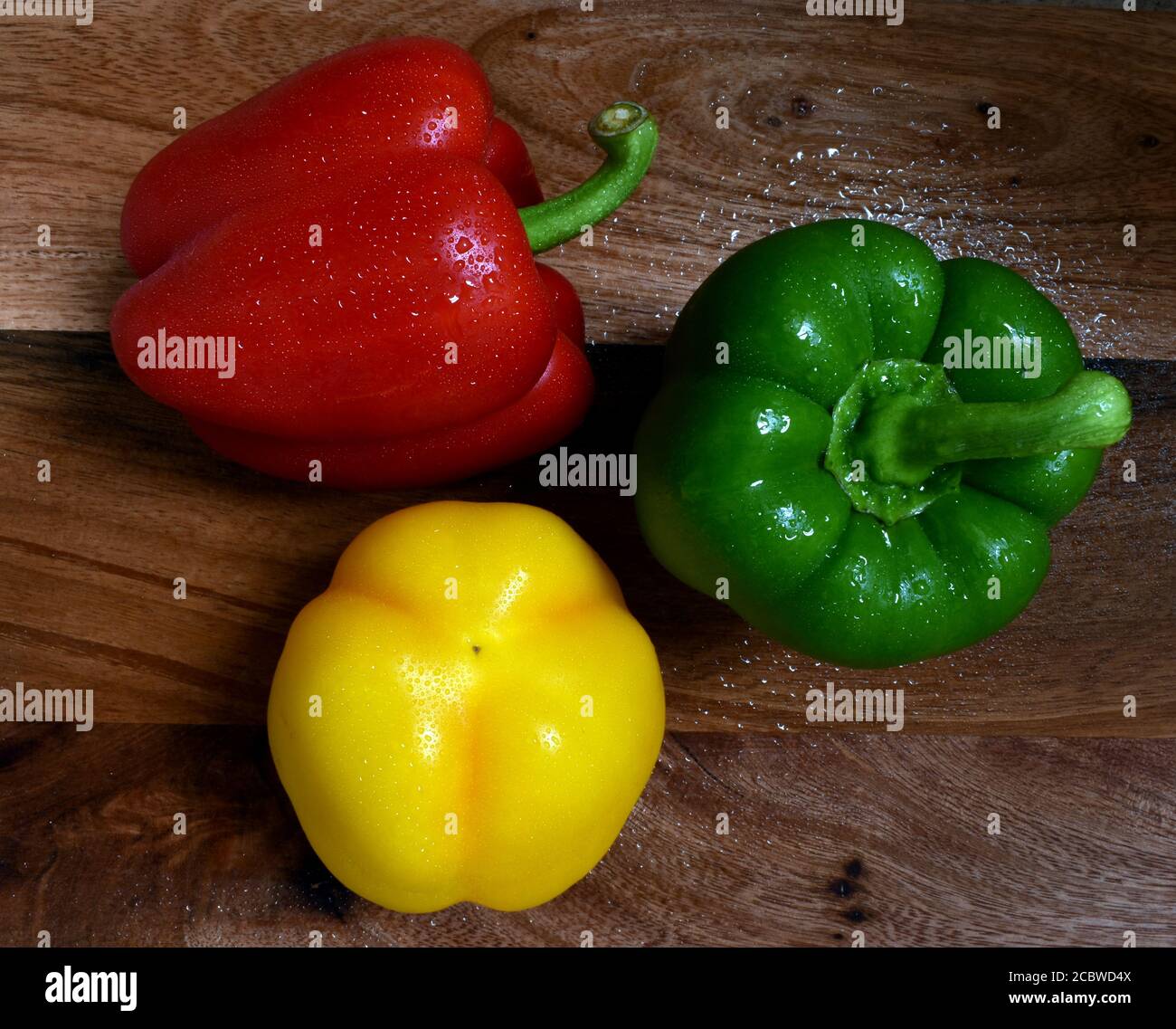 Sweet bell peppers (capsicum), Red green and yellow, fresh, washed and wet showing top, side and beneath on wooden chopping board under natural light. Stock Photo