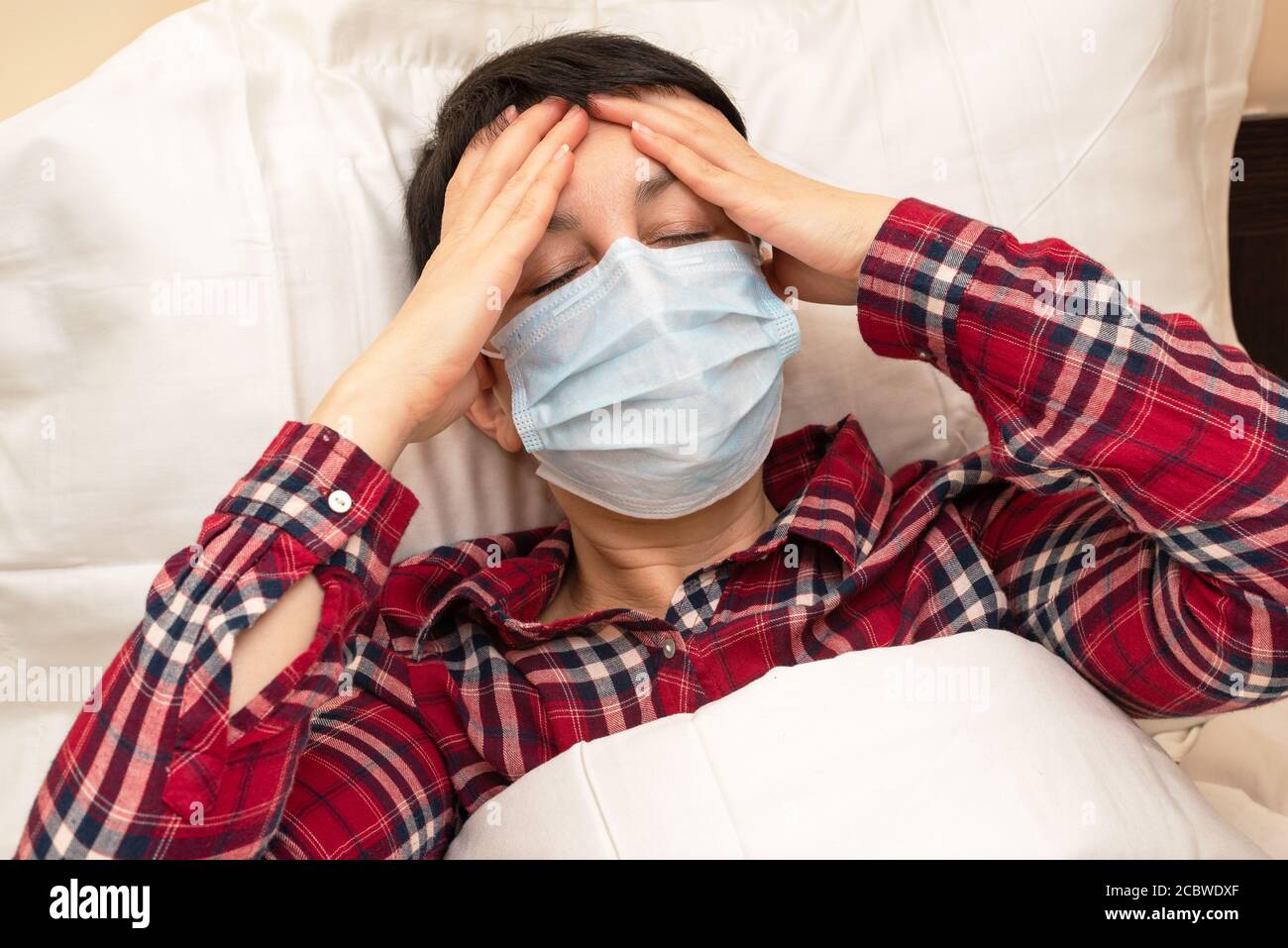 Sick woman in medical mask lying in bed suffering from fever and headache. Short-haired brunette with symptoms of covid-19 staying at home Stock Photo