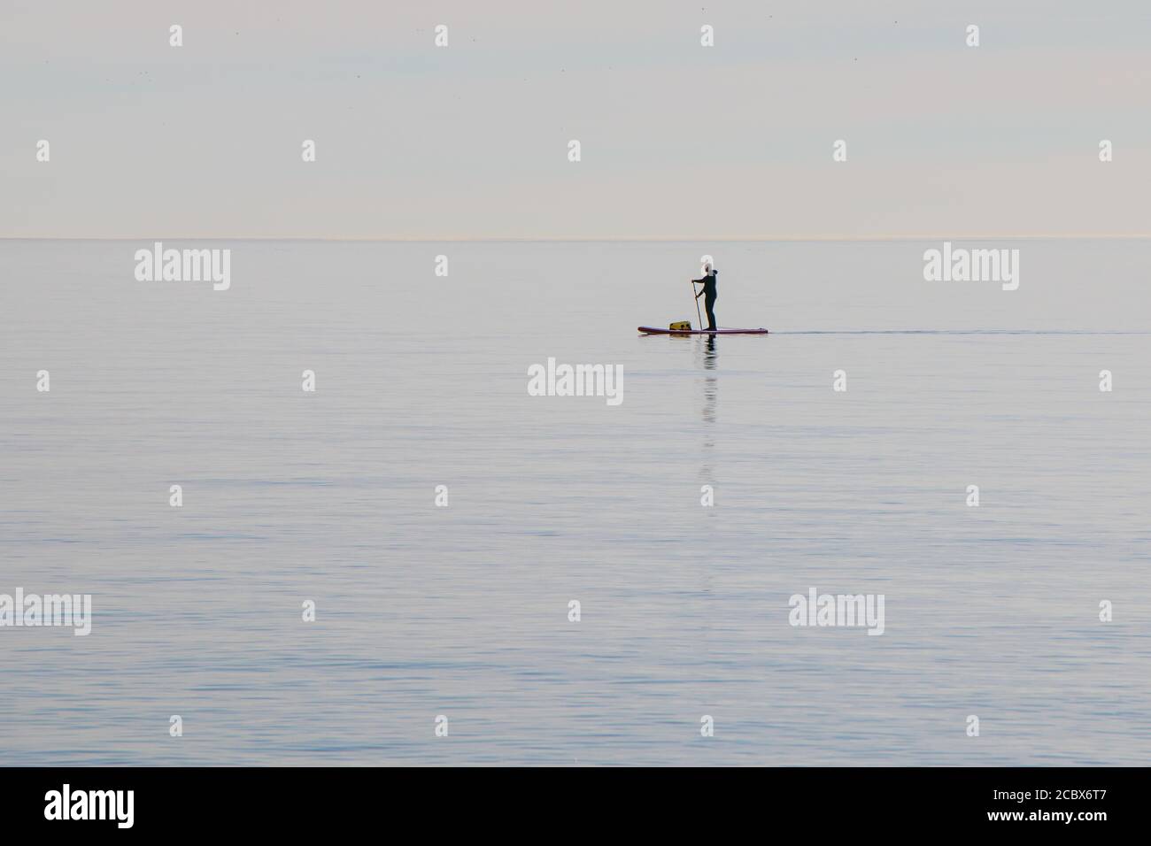 A lone man with a bag moving across the sea on a surfboard with an oar. Stock Photo