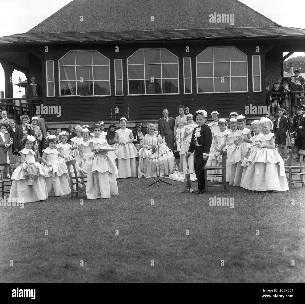 1950s, historical, the newly crowned Rose Queen sitting in her gown with other girls in their costumes and watched by local people in the playing field at Farnworth, Bolton, Lancashire, England, UK. Dating back to the 1880s, the annual Rose Queen festival, held in June, became a major annual event in many towns and villages across the UK, especially in Lancashire, known as the red rose county, following the Wars of the Roses (1455-87). Stock Photo