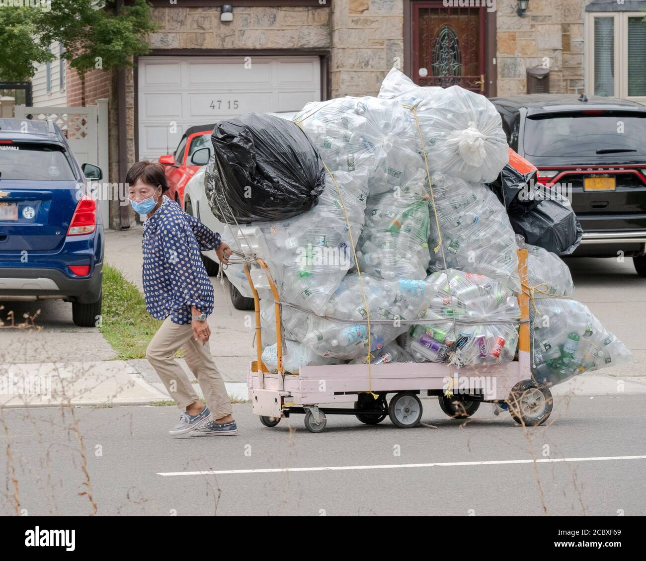 An Asian American, likely Chinese, woman drags a huge load of deposit bottles on a wheeled cart. On 162nd St. in Flushing, Queens, New York City. Stock Photo