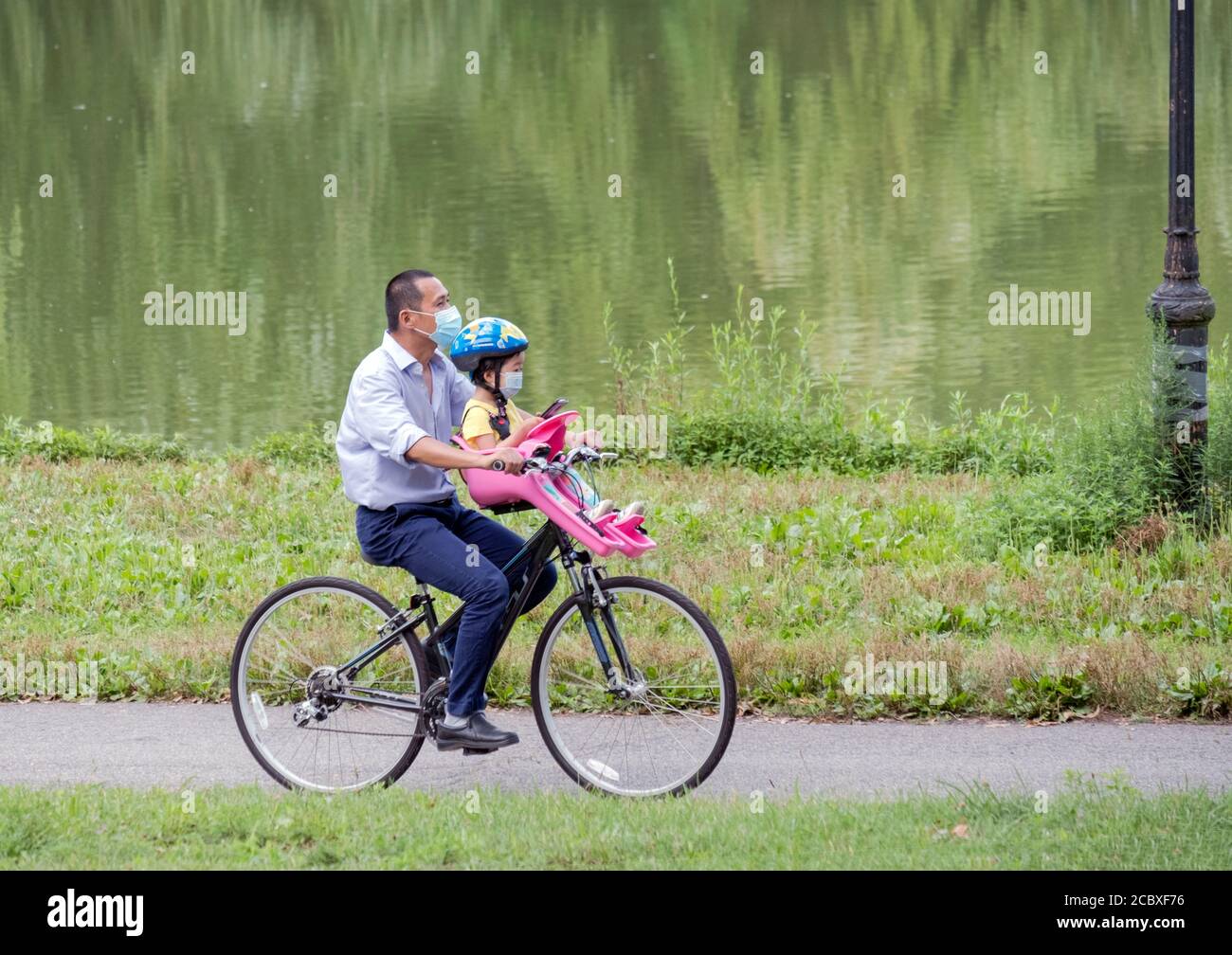 A father & his daughter, wearing masks, ride a bicycle equipped with a special kiddie seat. In Kissena Park in Flushing, Queens, New York Ci Stock Photo