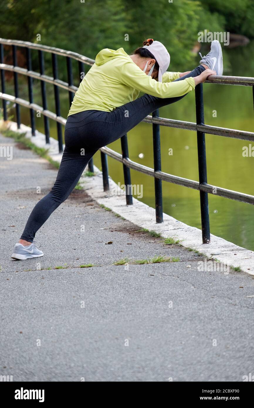 A nimble Asian American woman wearing a mask stretches before exercising. In Kissena Park, Flushing, New York City. Stock Photo