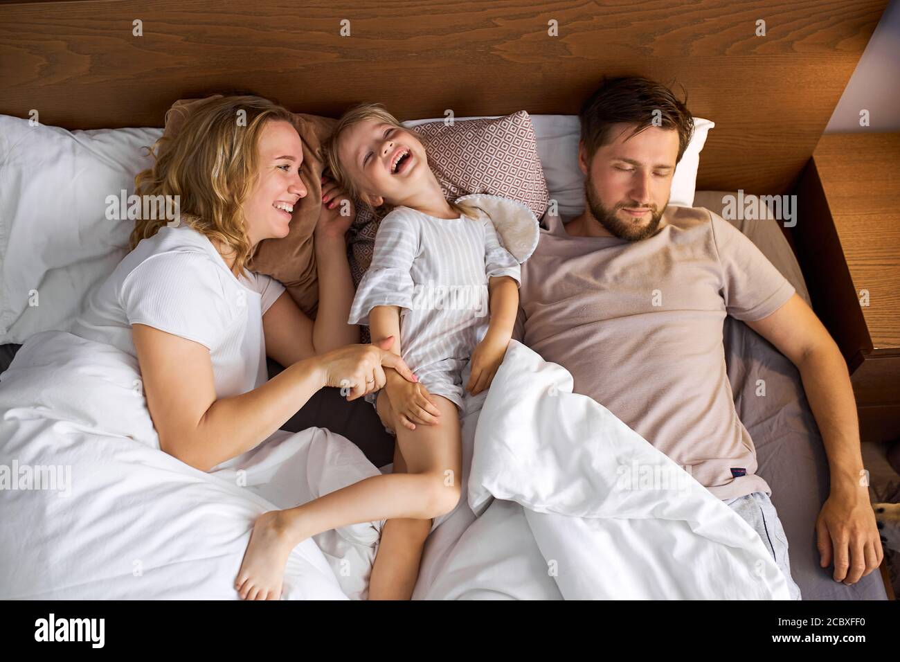 mother and daughter have fun on bed while father is sleeping, family on bed, weekends concept Stock Photo