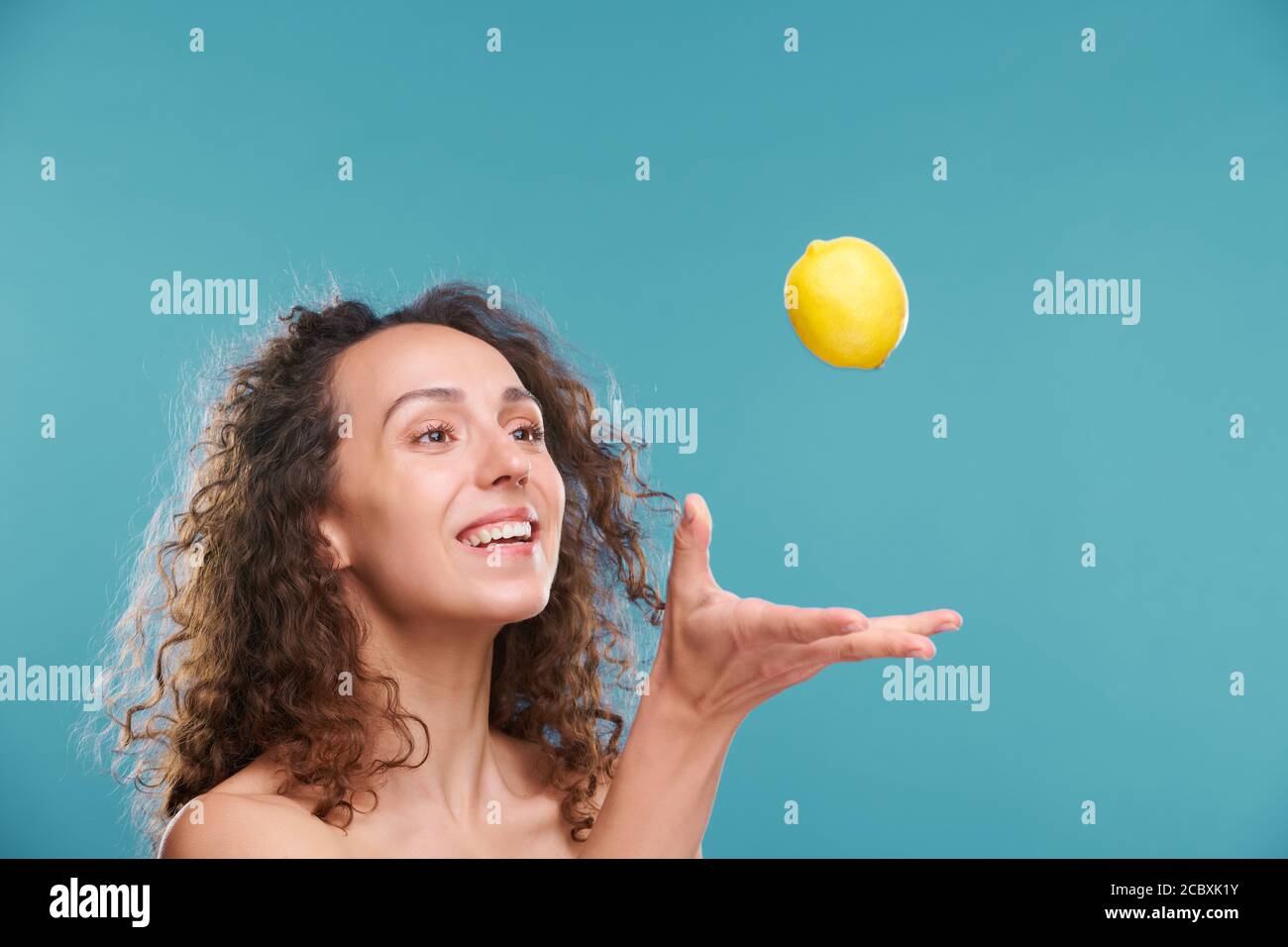 Young joyful woman with toothy smile and dark wavy hair throwing fresh lemon Stock Photo