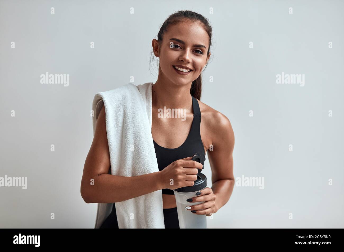 Positive smiling attractive woman in sportswear holding water bottle with towel on shoulders. Isolated in grey studio, room Stock Photo