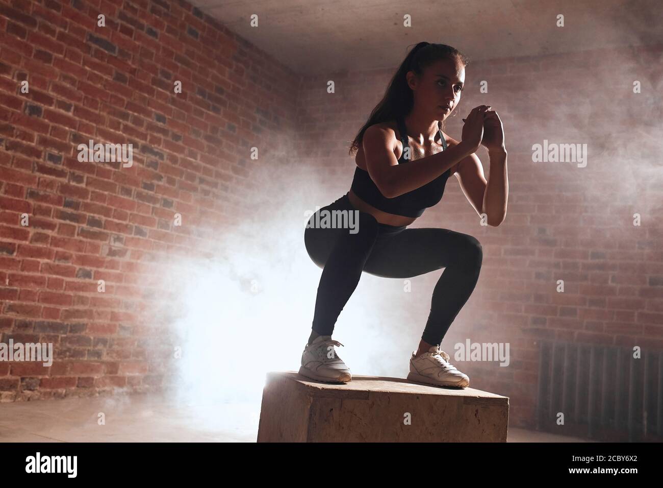 Slim woman jumping on wooden fit box, performing squats. Steam back