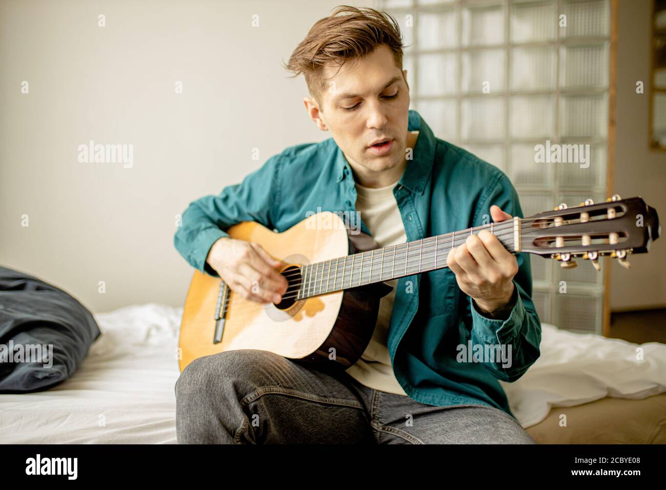 young caucasian guy play acoustic guitar at home, man keen on music, perform music. handsome male in casual wear sit on bed with guitar. indoors Stock Photo
