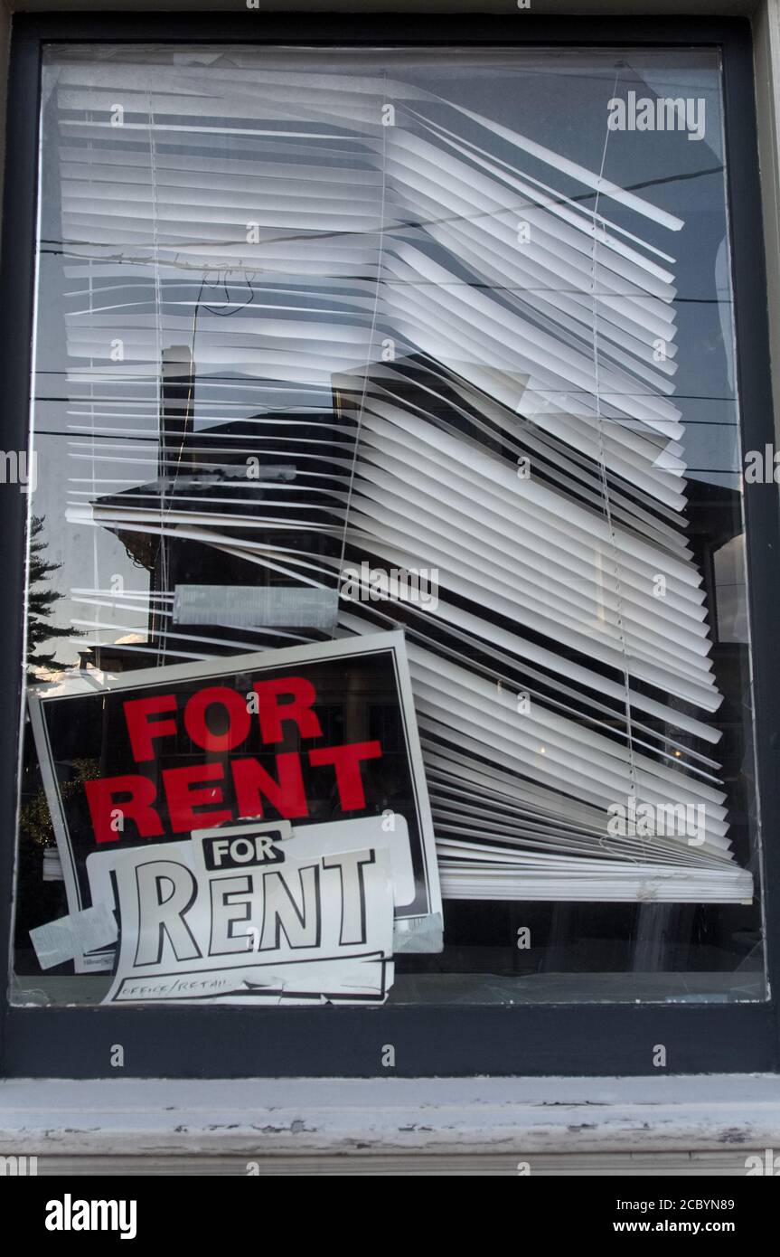 A crooked 'For Rent' sign in the corner of a window, with dilapidated blinds Stock Photo