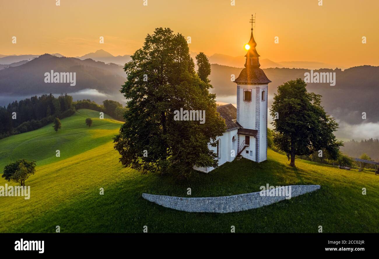 Skofja Loka, Slovenia - Aerial view of the beautiful hilltop church of Sveti Tomaz (Saint Thomas) with amazing golden foggy sunrise and the Julian Alp Stock Photo