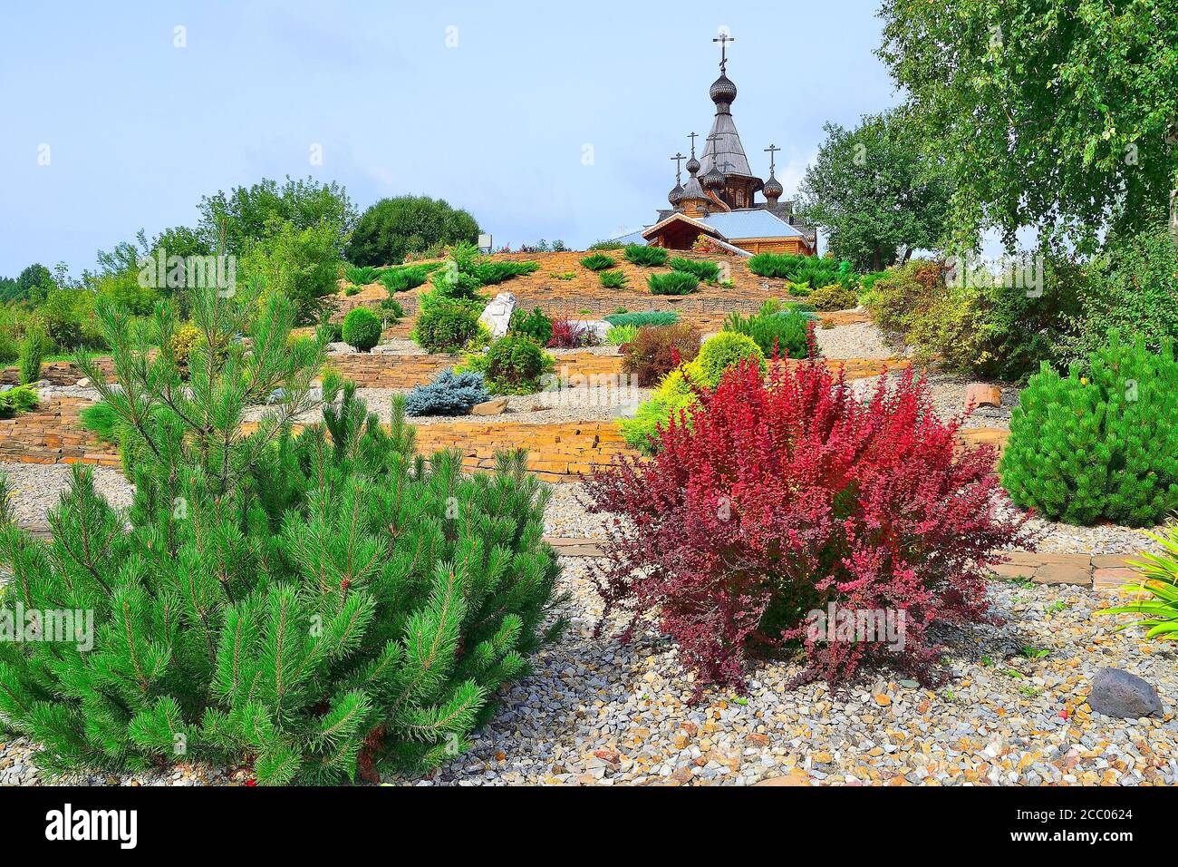 Dwarf trees, bushes and flowers on the stony terraces fortified by gabions on hill slope - front yard of orthodox Temple. Domes and crosses of church Stock Photo