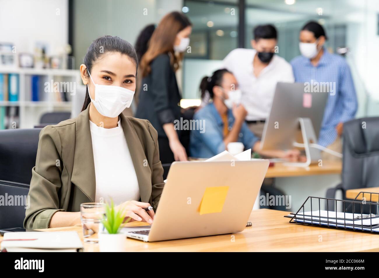 Portrait of asian office employee businesswoman wear protective face mask work in new normal office with interracial team in background as social dist Stock Photo