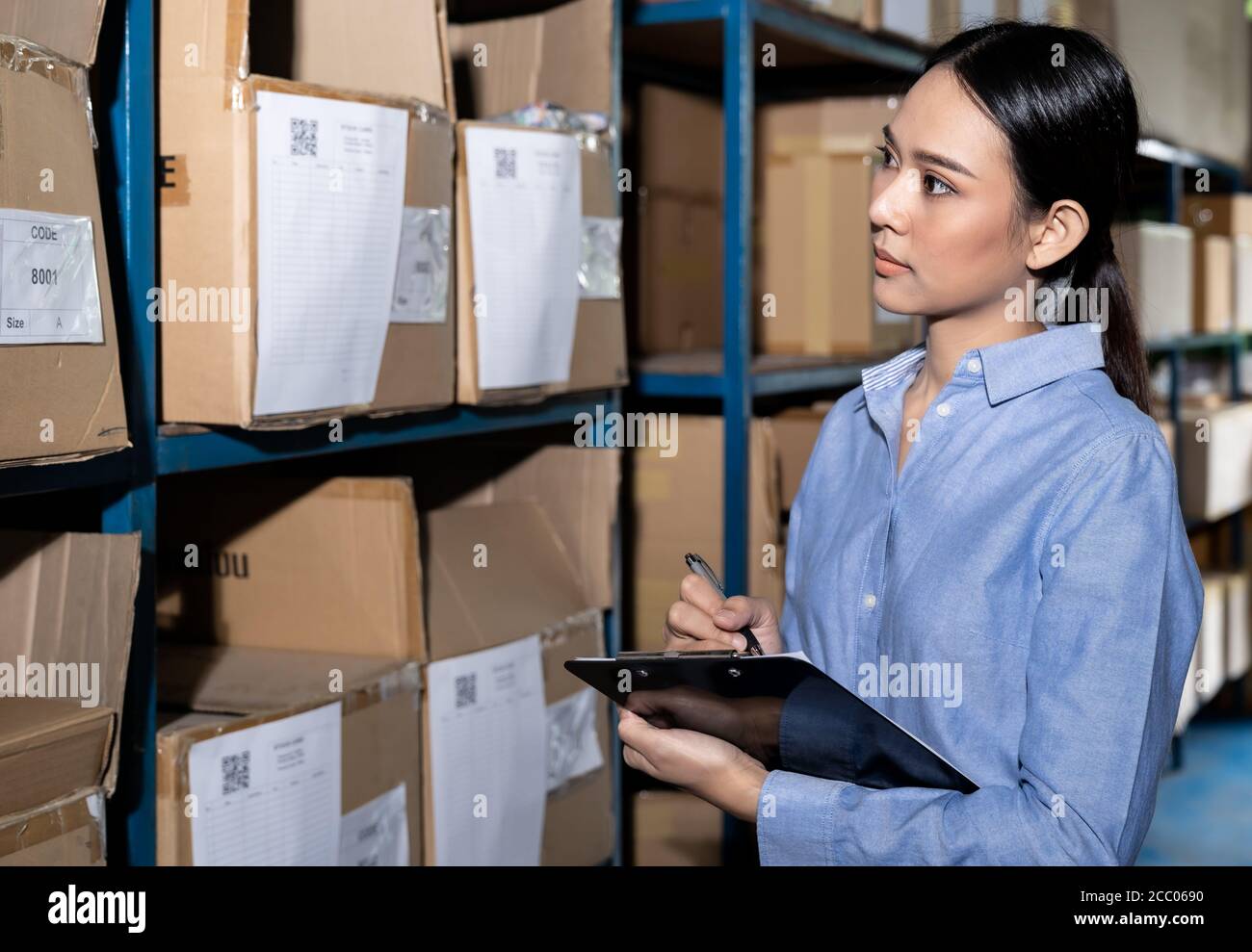 Close up of Asian smart businesswoman working with product inventory and writing on clipboard in distribution warehouse. For business warehouse invent Stock Photo