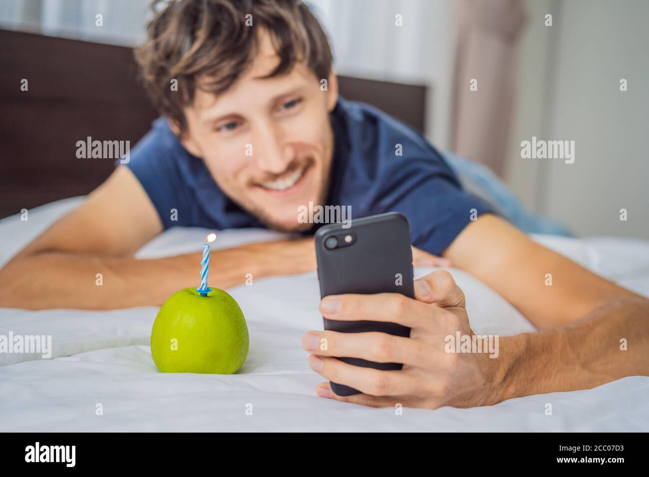 Quarantine birthday. Man with a smartphone and birthday cake-apple with candle. Stay home while quarantine. Online call. Video conference Stock Photo